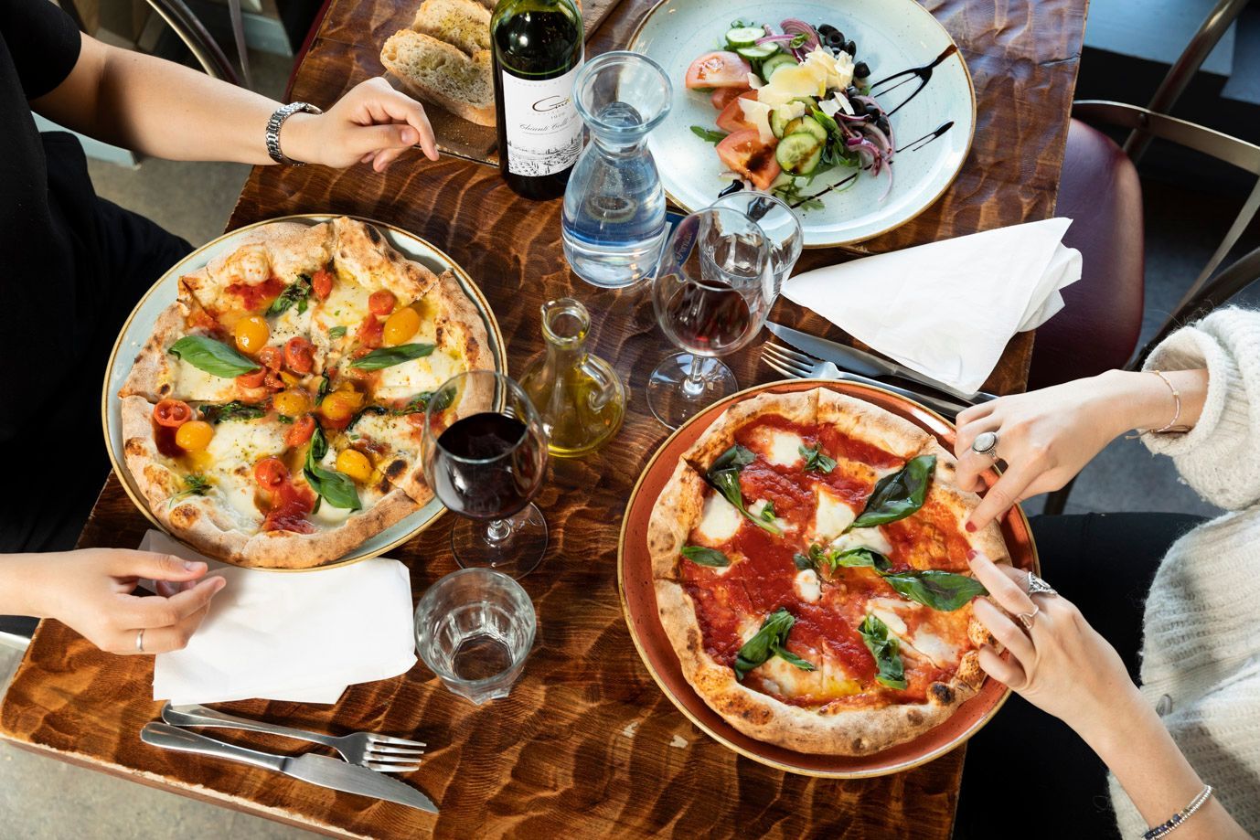 Two pizzas, salad, and wine on a table with two people's hands reaching for the food.