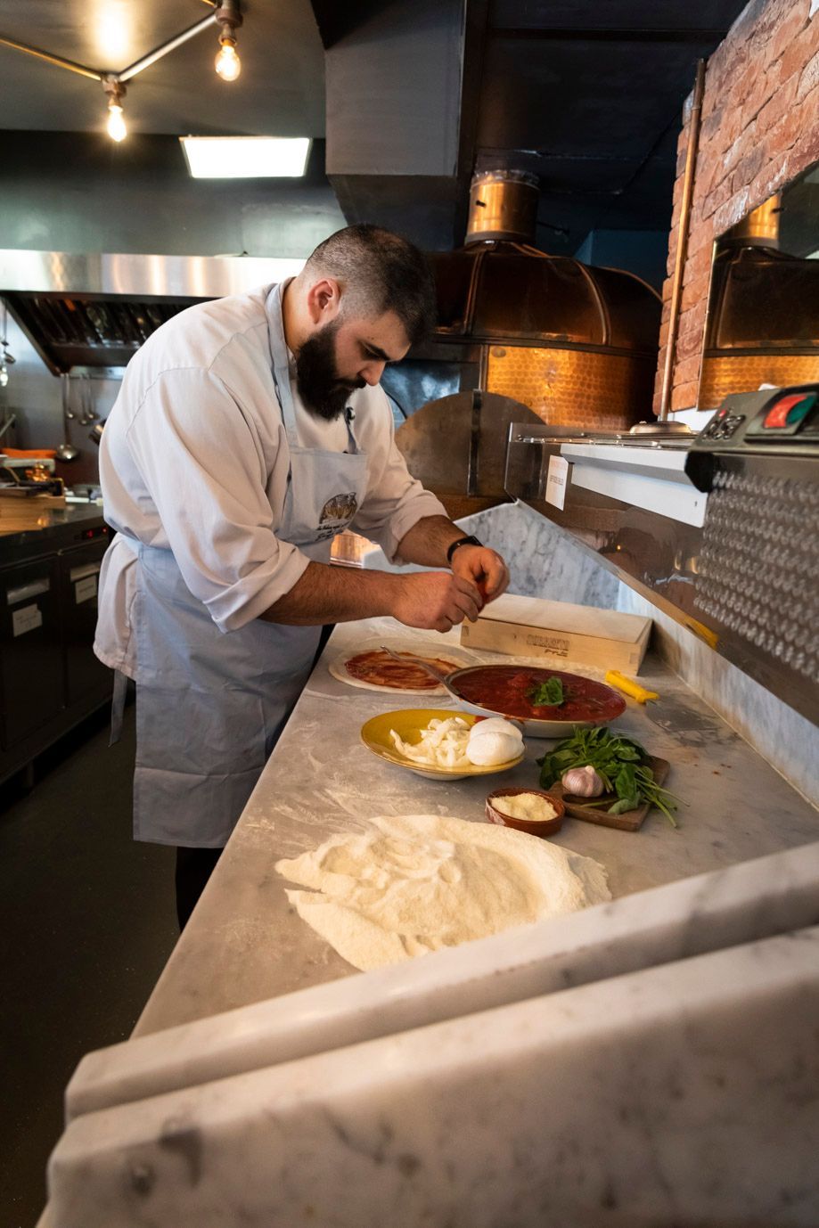 Chef in apron prepares pizza with toppings in a restaurant kitchen.