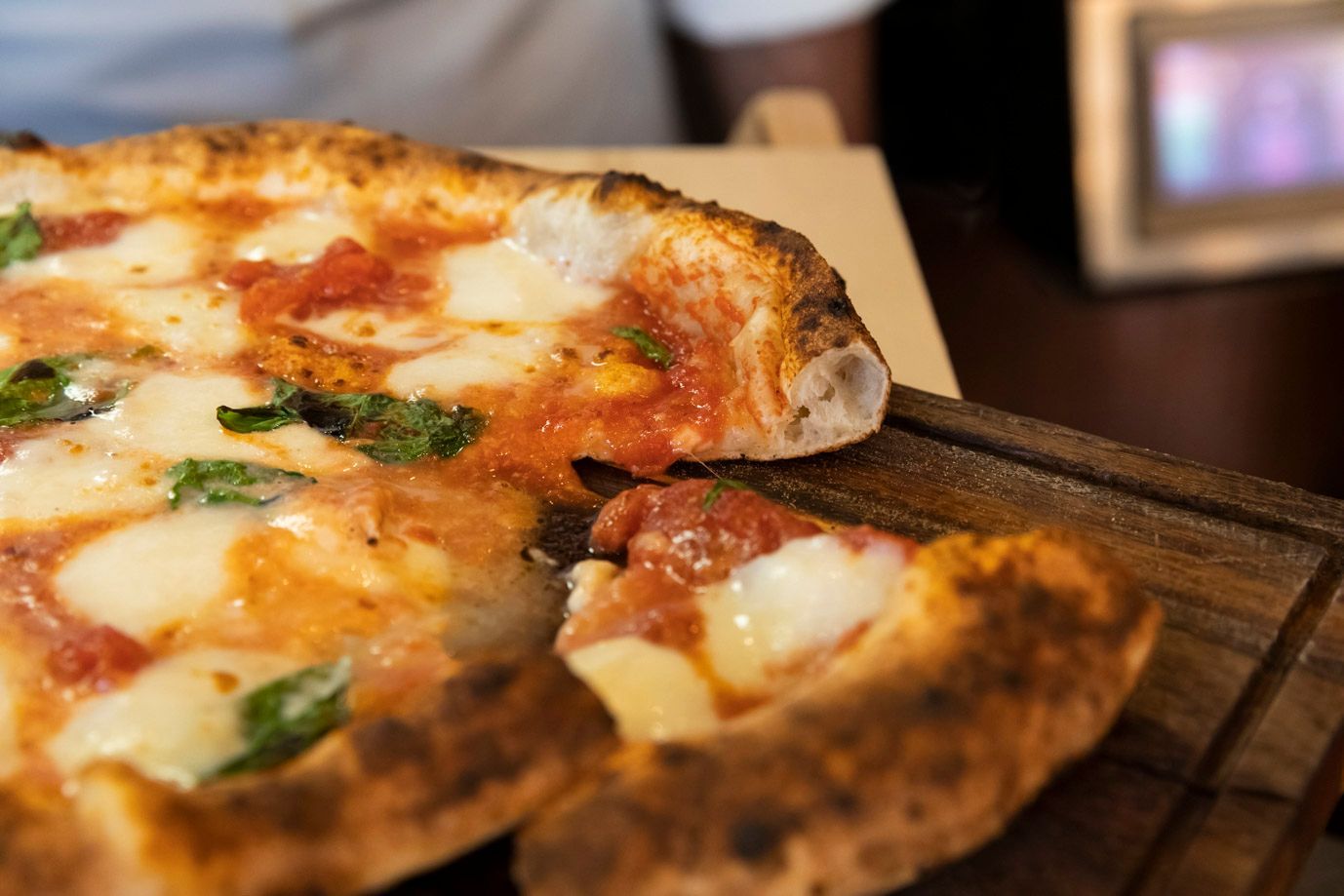 Pizza slice being lifted from a wood board. Cheese, tomato sauce, and basil visible. Close-up view.
