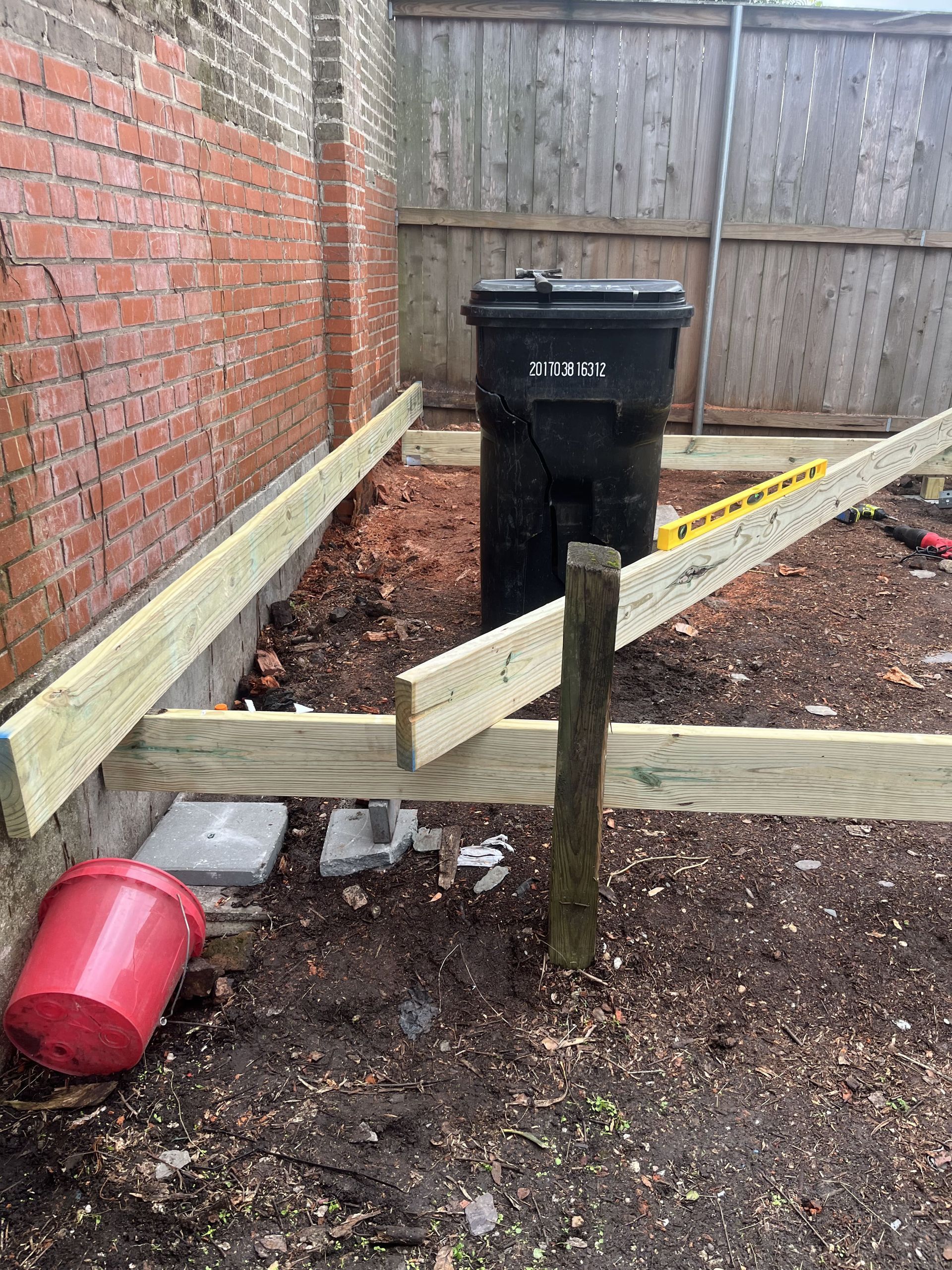 A trash can is sitting next to a wooden post in a backyard.