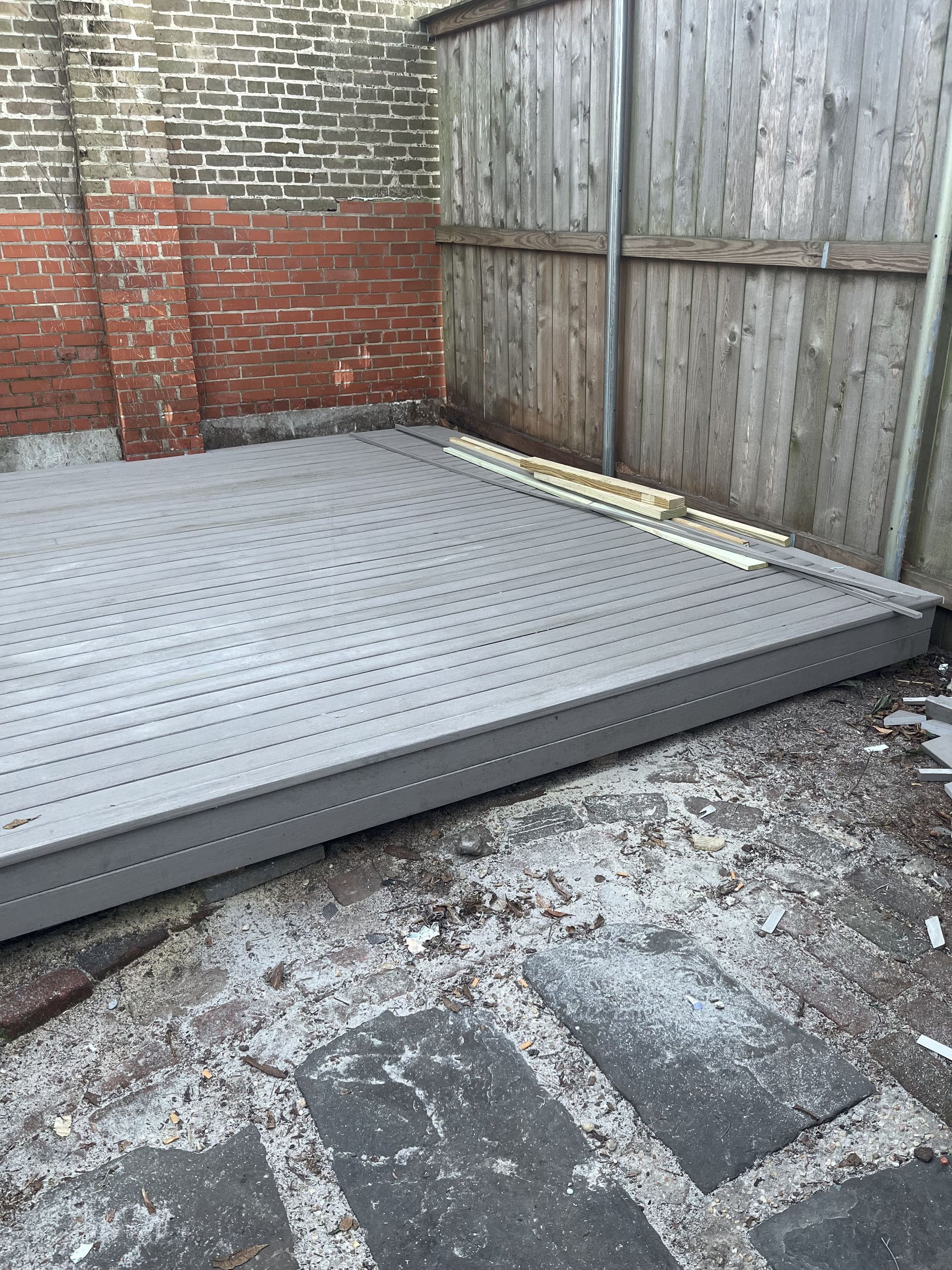 A wooden deck is sitting in a backyard next to a brick wall and a wooden fence.