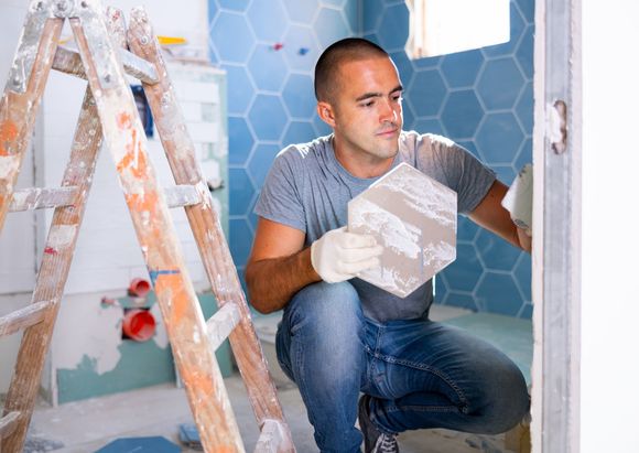 A man is kneeling down in a bathroom holding a tile.