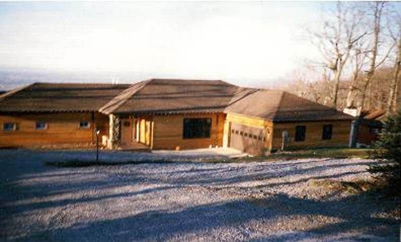 Brown log cabin-style building with a garage, set on a gravel driveway, against a backdrop of mountains and bare trees.