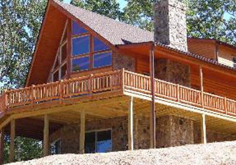 Wooden cabin with stone chimney, wraparound deck, and large triangular window.