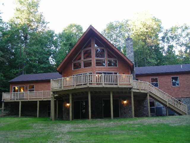 Wooden house with large deck, a-frame roof, and stone chimney, set in a wooded area.