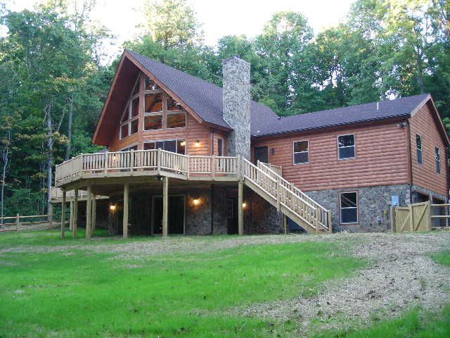 Wooden house with a large deck and stone chimney, set in a green grassy area surrounded by trees.