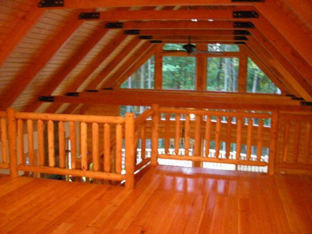 Loft interior with wooden railing, beams, and hardwood floor; view to windows and trees.