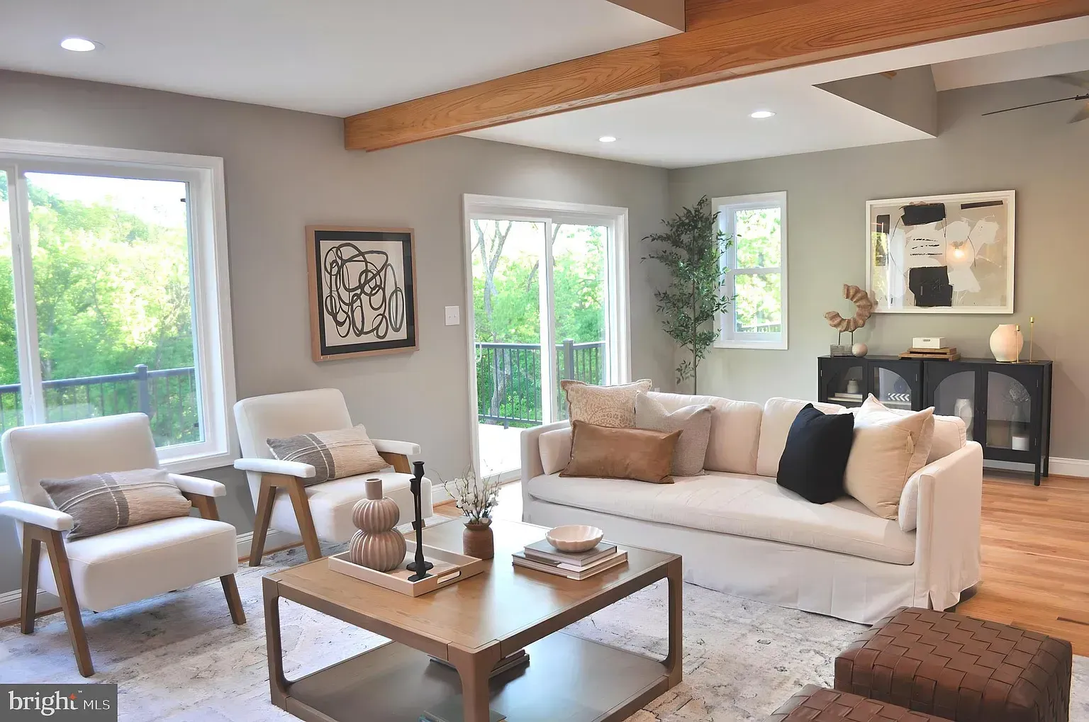 Living room with cream-colored furniture, wooden beams, large windows, and art.