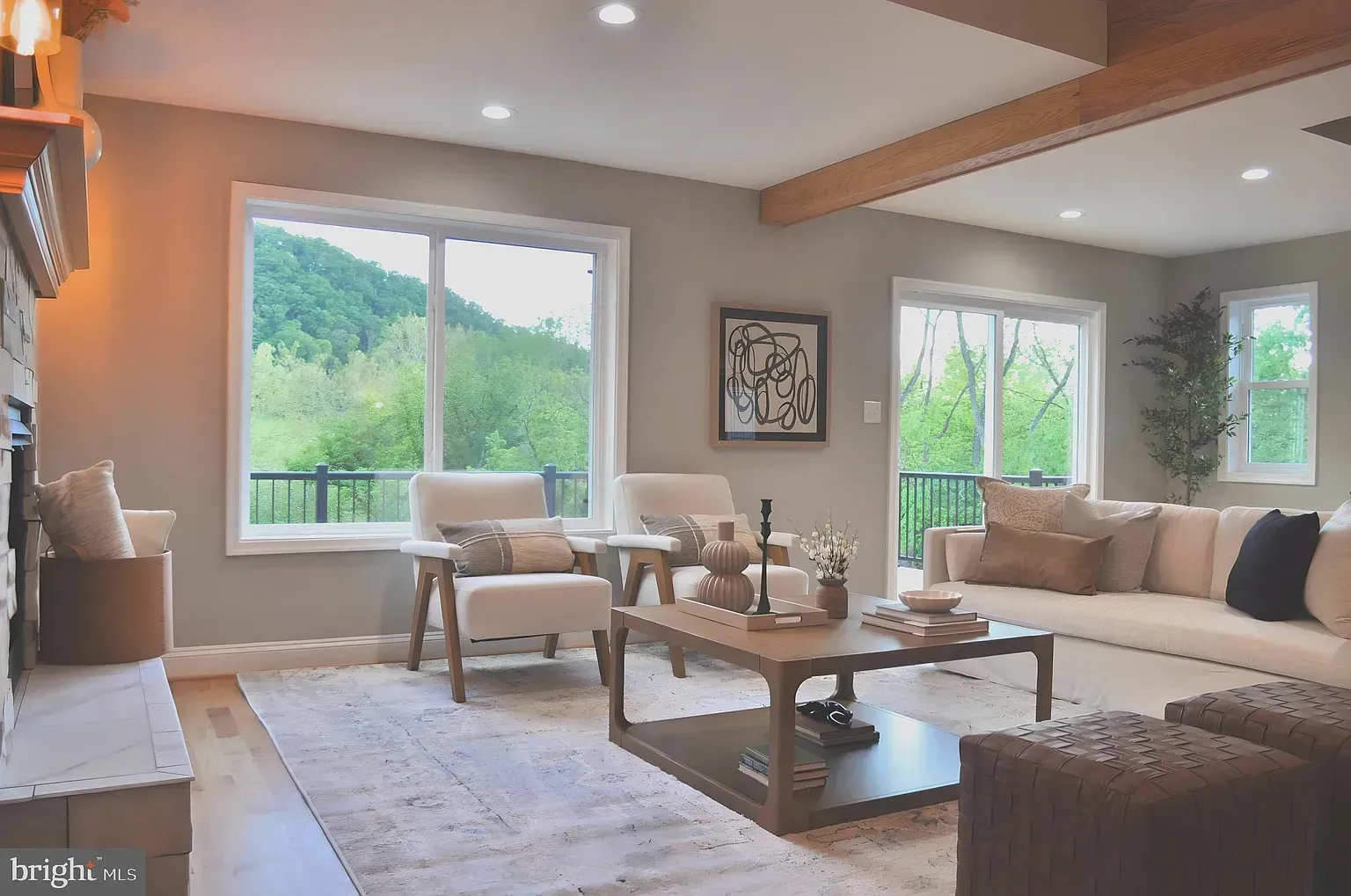 Living room with large windows overlooking a green landscape. Beige sofa, chairs, and wooden coffee table.