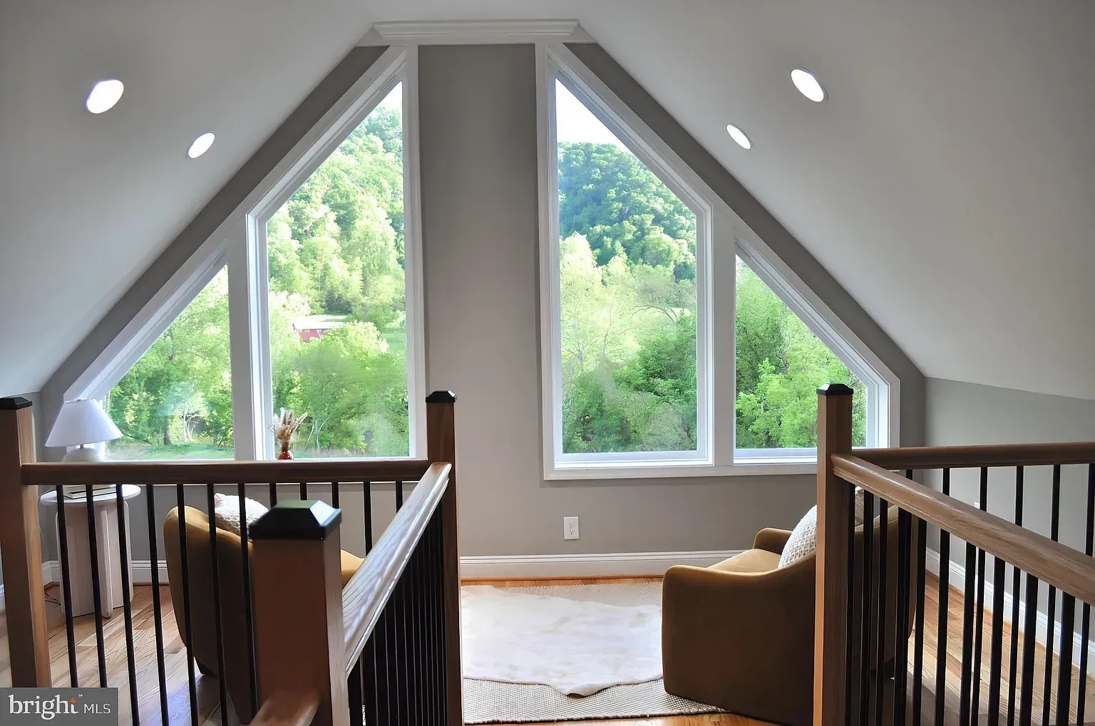 Loft with A-frame windows overlooking greenery; chairs, wood railing, and recessed lights.