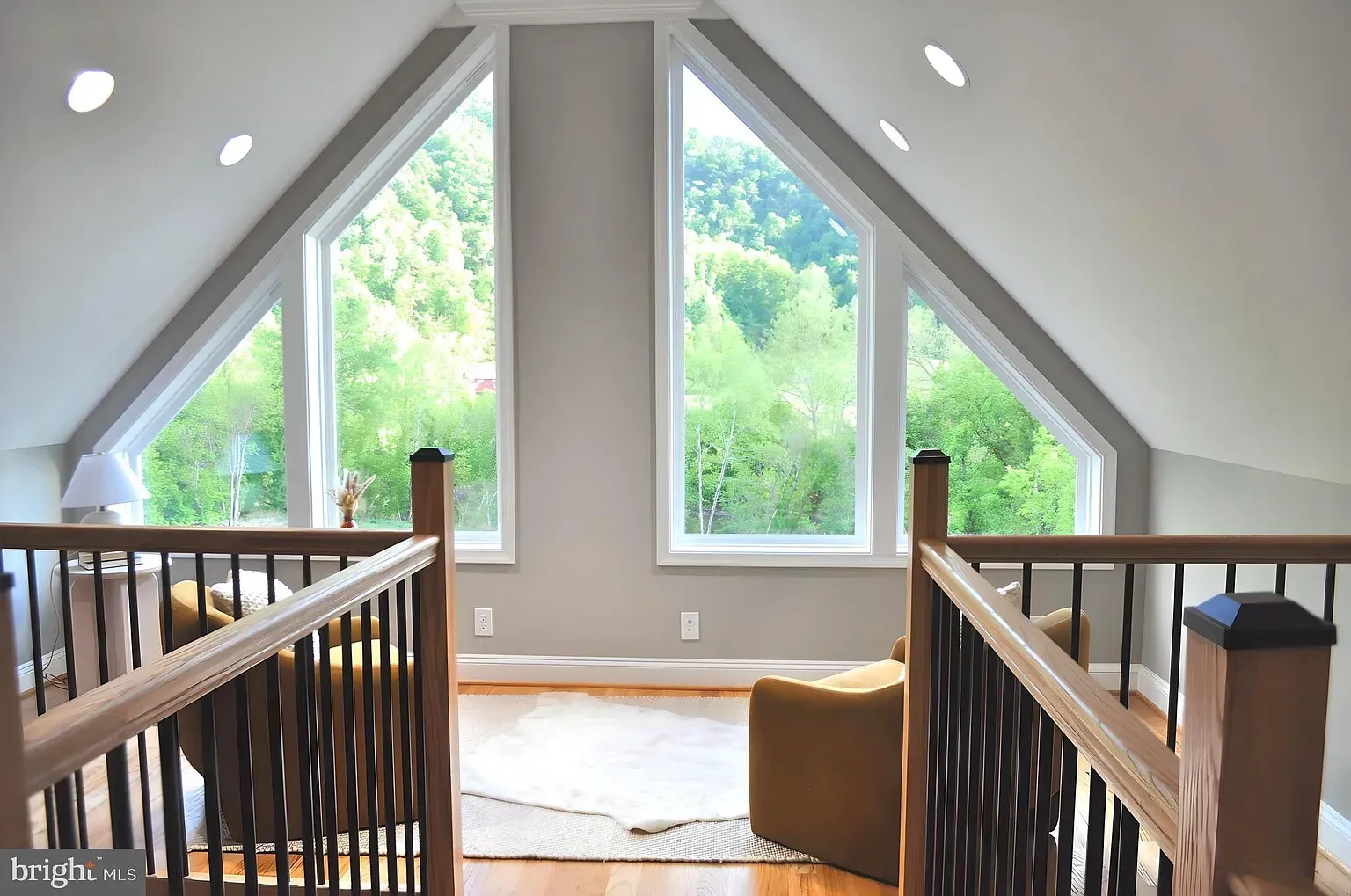 Loft with angled windows, wooden railing, and seating area overlooking greenery.