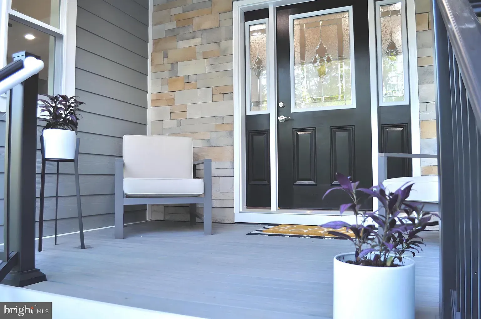 Front porch with black door, stone accent, grey siding, potted plants, and a chair.
