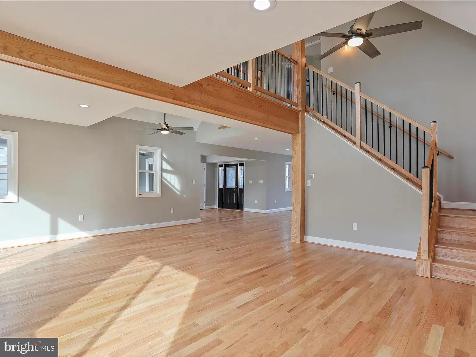 Spacious interior with hardwood floors, stairs, and a balcony. Light grey walls, wooden beams, and two ceiling fans.