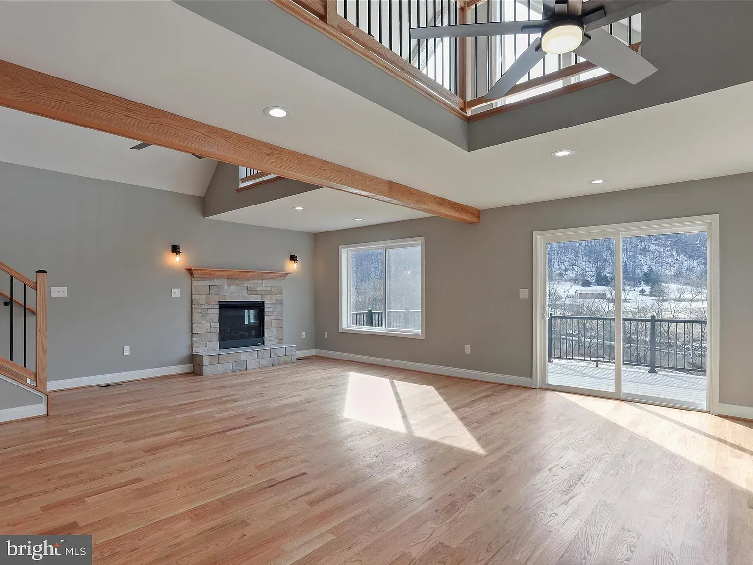 Spacious living room with wood floors, stone fireplace, and deck access. Gray walls, exposed beams, and high ceilings.