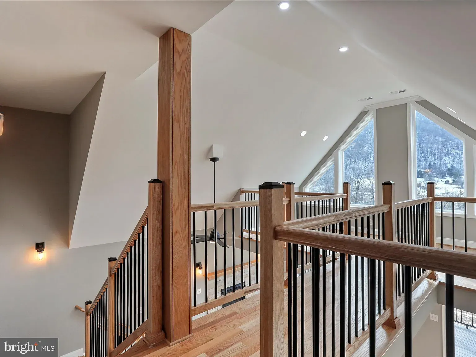 Interior view of a staircase with a wooden post and black railing, leading to a loft with large windows.
