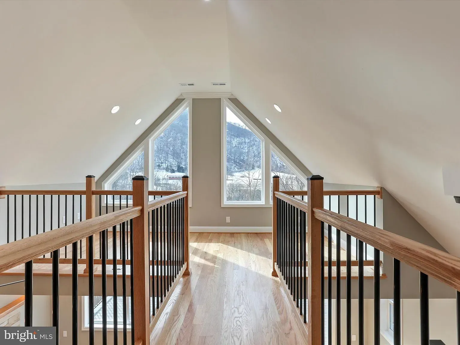 Interior loft with a wood floor and railing, overlooking mountain views through triangular windows.