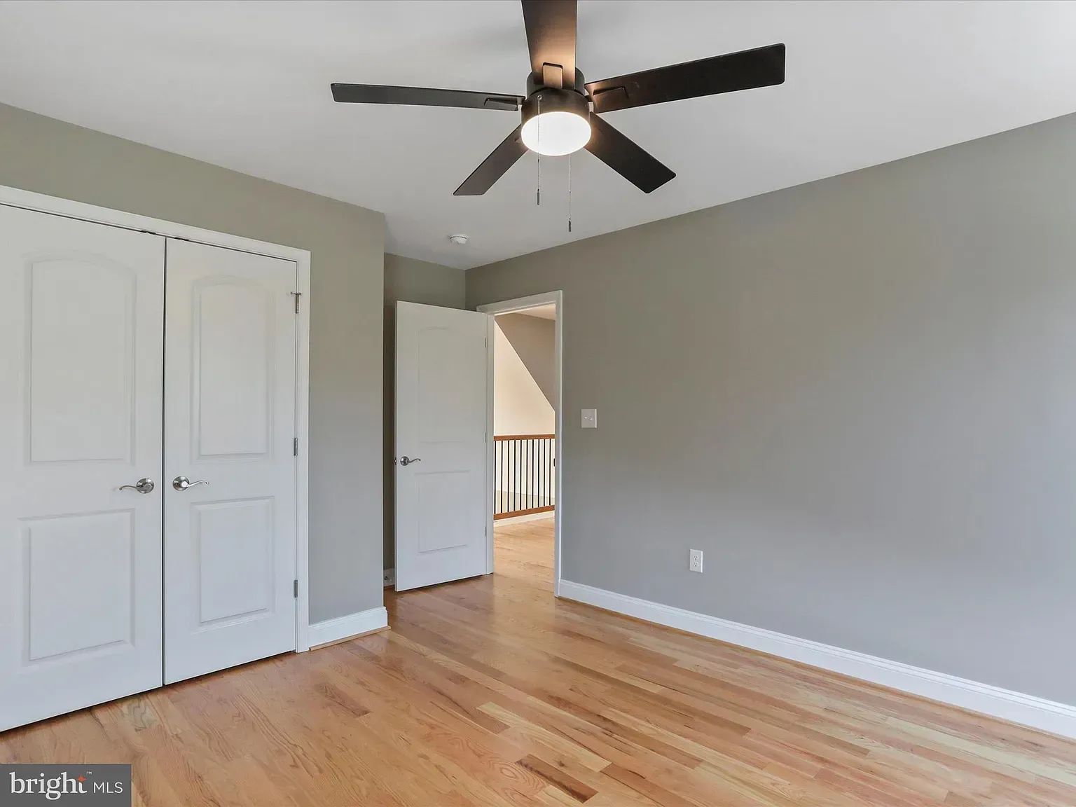 Bedroom with hardwood floors, gray walls, white closet doors, and ceiling fan. Open doorway leads to staircase.