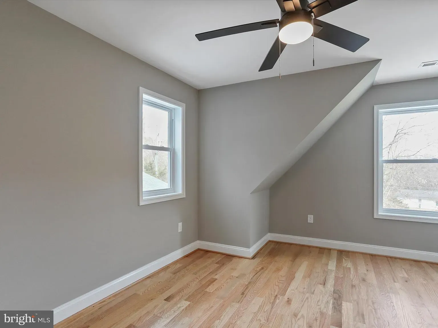 Empty room with hardwood floors, two windows, and a ceiling fan; gray walls and an angled ceiling.