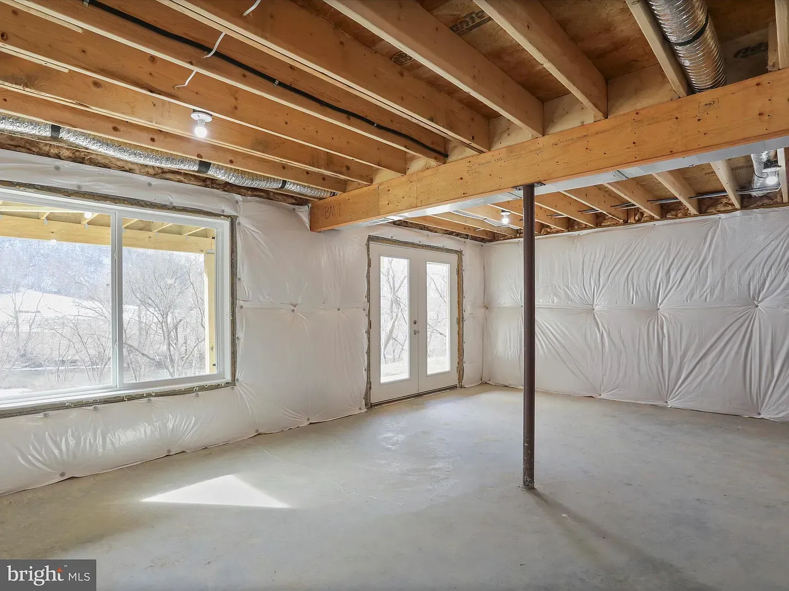 Unfinished basement with concrete floor, white insulation, and wood beams. French doors and large window.