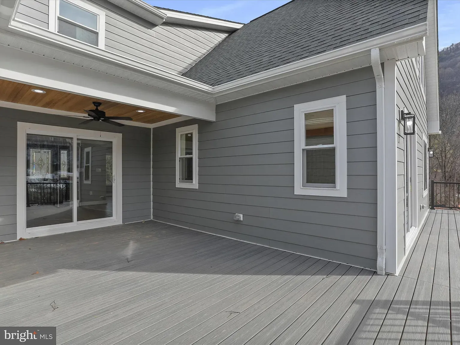 Gray house exterior with deck. Sliding glass doors, windows, and light gray siding. Sunny day.