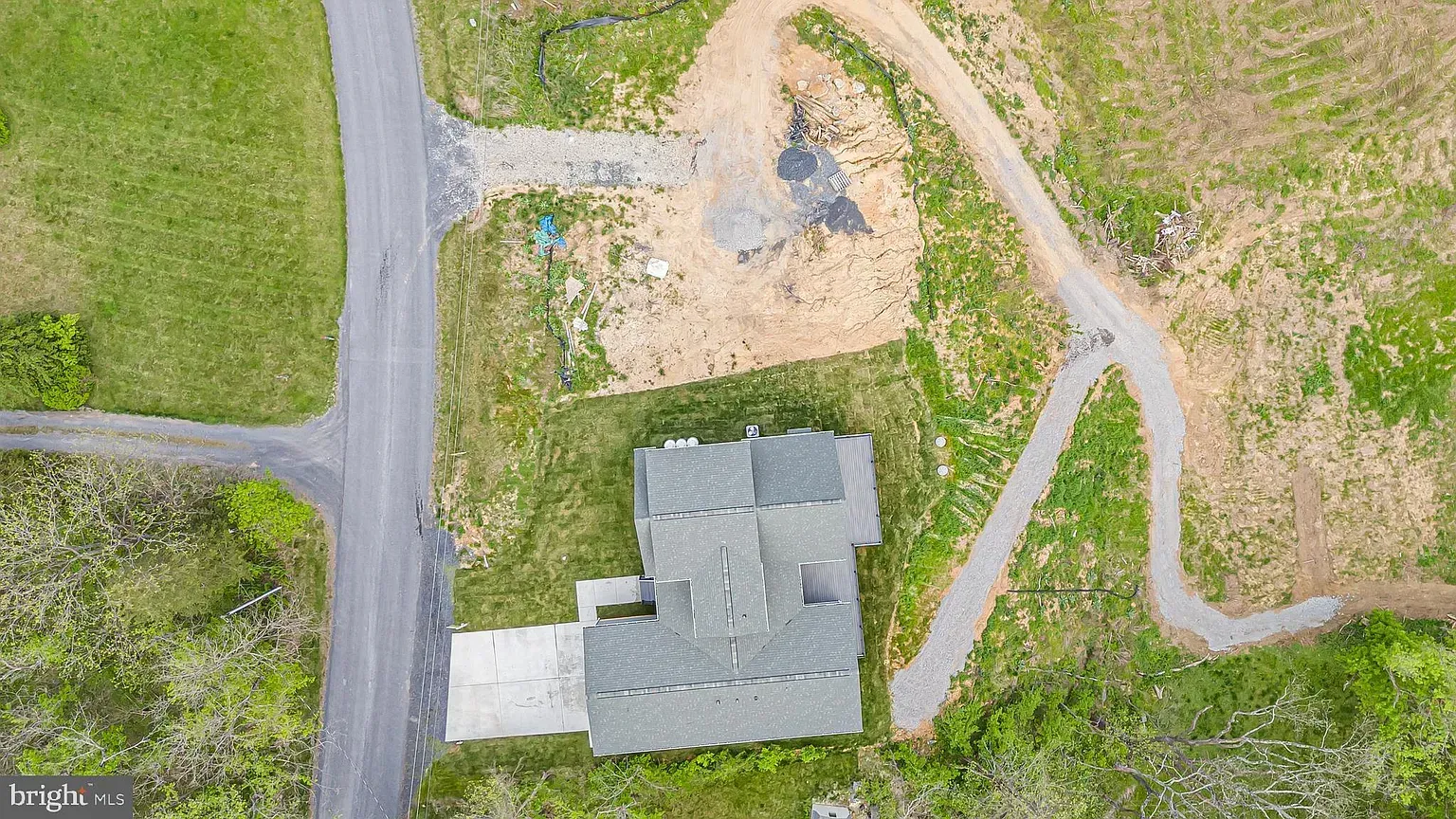 Aerial view of a house with a gray roof and driveway, surrounded by grassy areas and dirt paths.