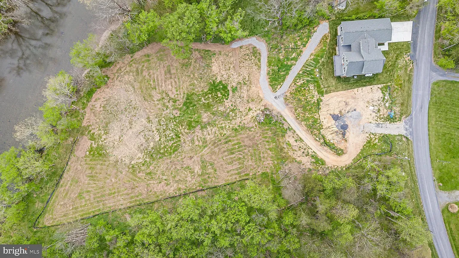 Aerial view of a cleared lot and a house with a driveway next to a road and a body of water.