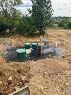 Green septic tanks and related equipment in an excavated area, surrounded by dirt and vegetation.