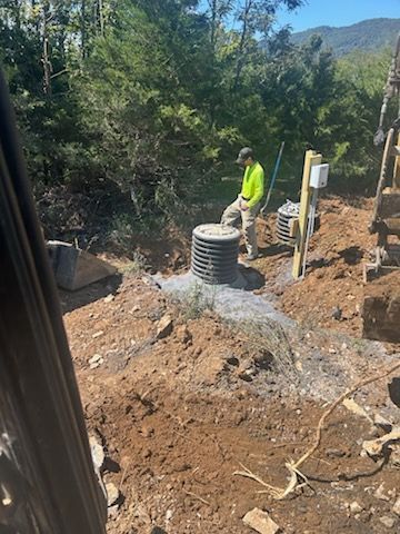 Construction worker near a gray septic tank, other equipment, and forest in background.