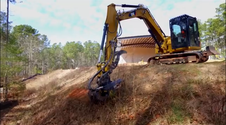 Yellow excavator on a hillside, clearing brush. Sunlight, trees, and a building in the background.