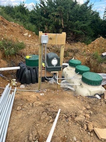 Septic system components installed outdoors: tank, pump, and control box, surrounded by earth.