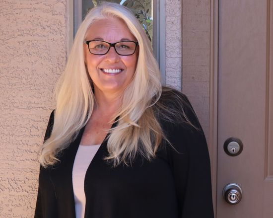 Woman with glasses smiles in front of a tan wall and brown door.