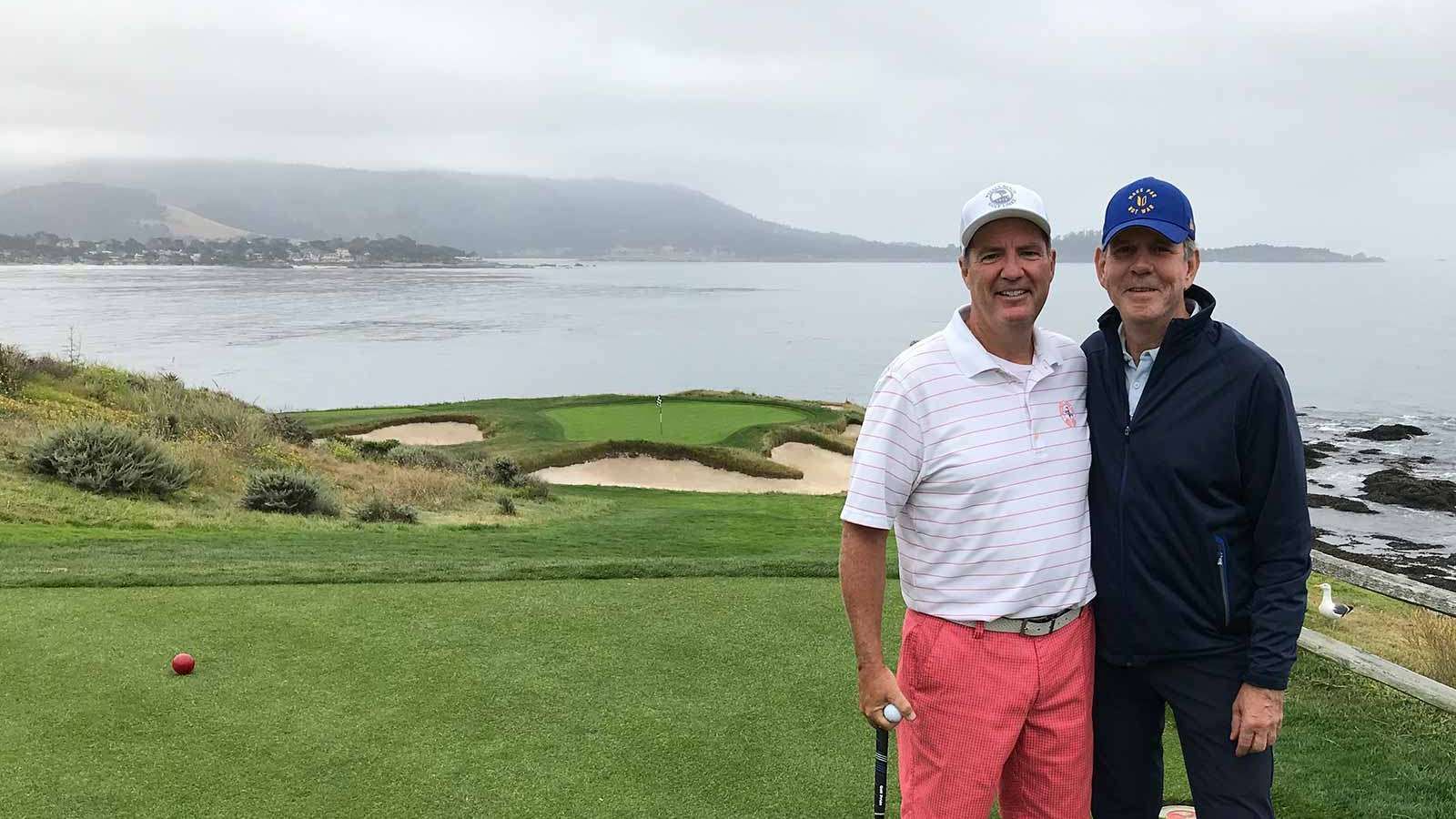 Two men on a golf course overlooking the ocean, one with a red golf tee.