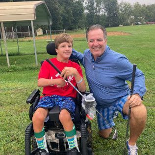 A man is kneeling next to a boy in a wheelchair holding a golf club.