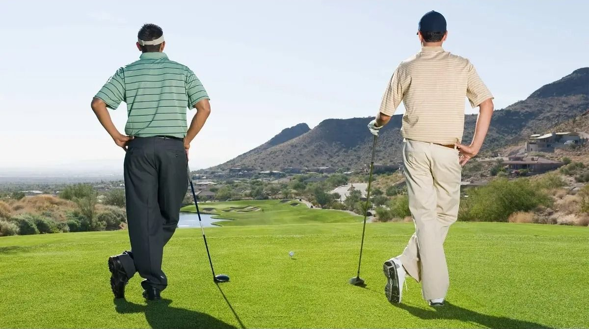 Two golfers on a green tee, overlooking a scenic course, hands on hips, backs to the camera.