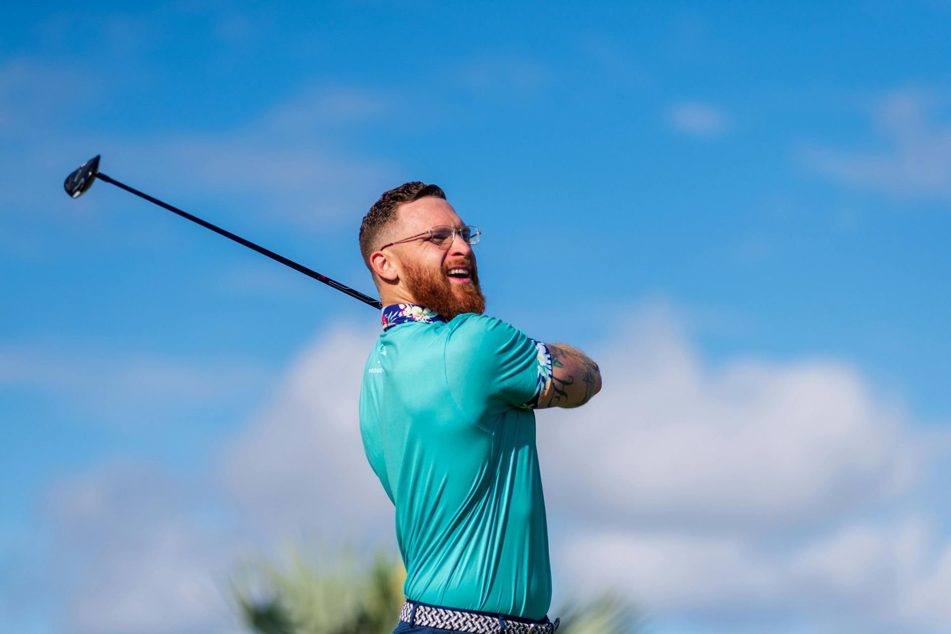 Man with red beard swinging a golf club on a sunny golf course, wearing teal shirt and glasses.