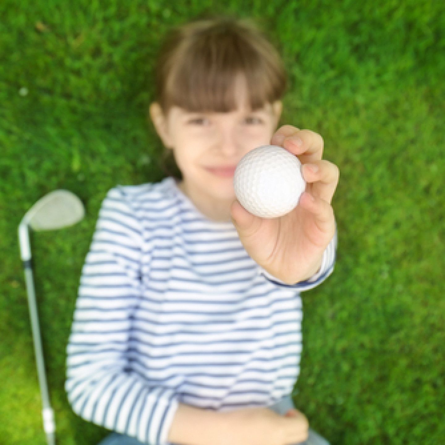 A young girl is laying on the grass holding a golf ball.