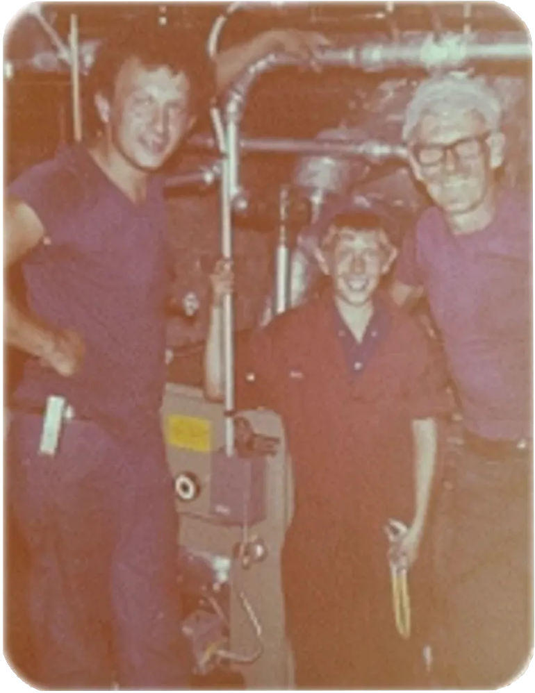 Three people in work uniforms stand near equipment. Two smile, and one holds a tool. The setting is indoors.