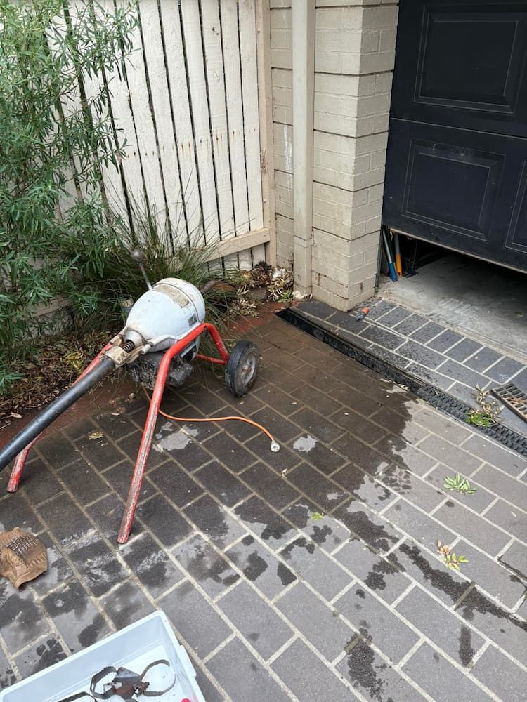 A Red and Silver Drain Cleaning Machine Sits on Wet Brick Pavers Near a Garage — AD Plumbing & Maintenance in Nicholls, ACT