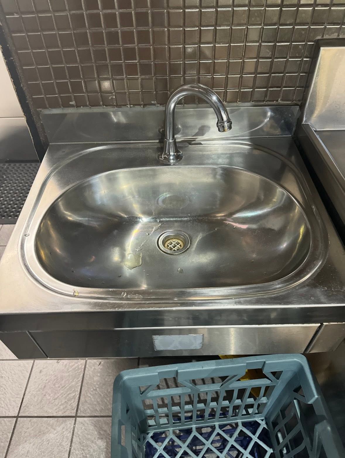 Stainless steel sink with faucet and drain, set against tiled backsplash, with a blue crate below — AD Plumbing & Maintenance in Canberra, ACT