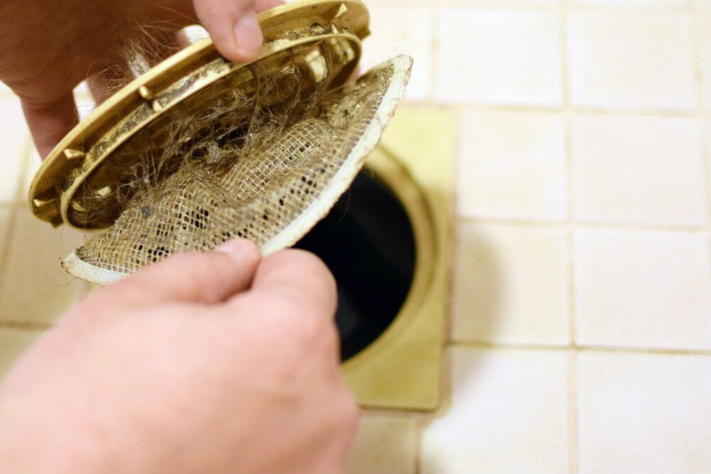 Hands Holding Up a Clogged Shower Drain Cover Over a Dark Drain Opening — AD Plumbing & Maintenance in Nicholls, ACT