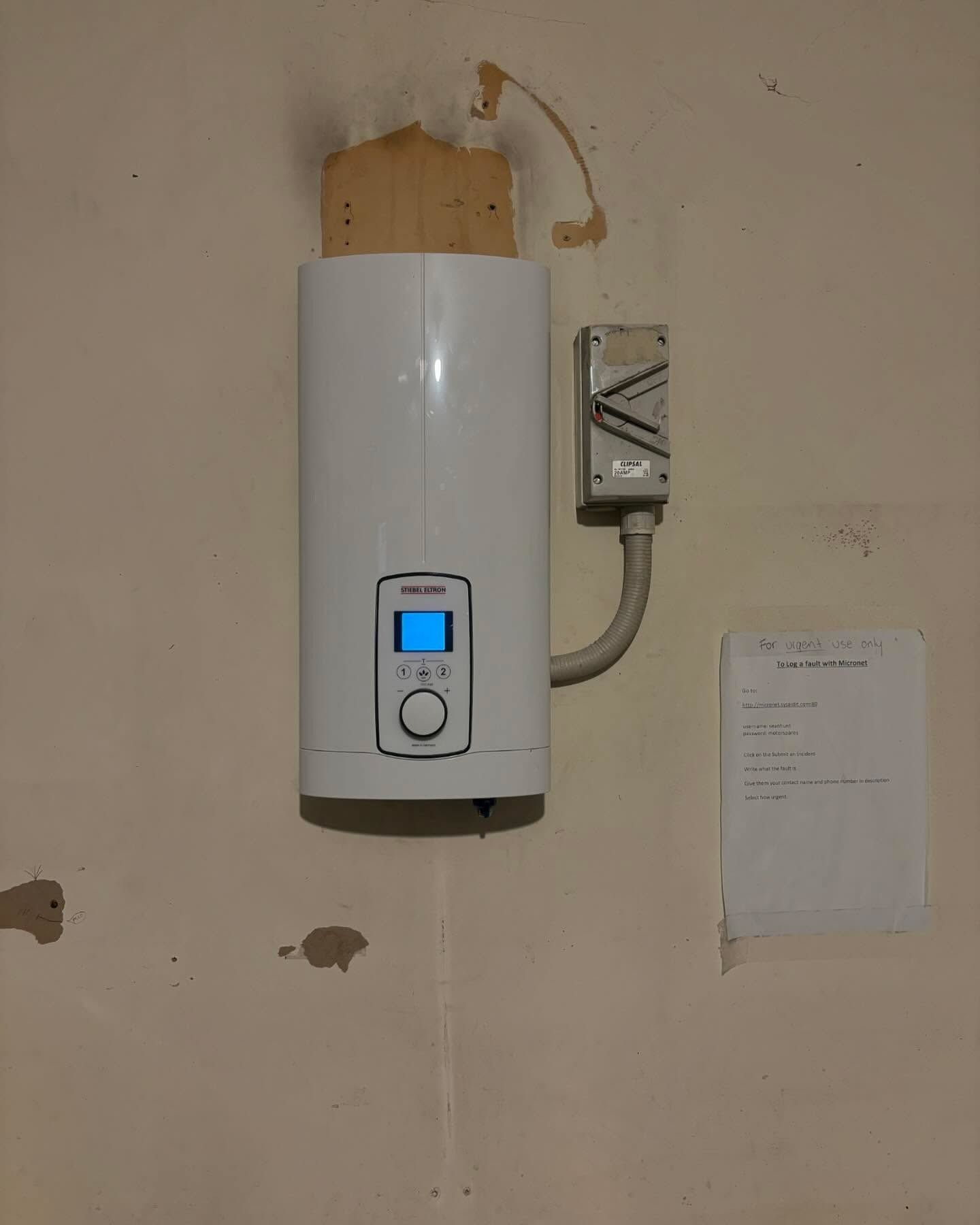 Wall-mounted Water Heater With Blue Display and Electrical Box on a Beige Wall — AD Plumbing & Maintenance in Canberra, ACT