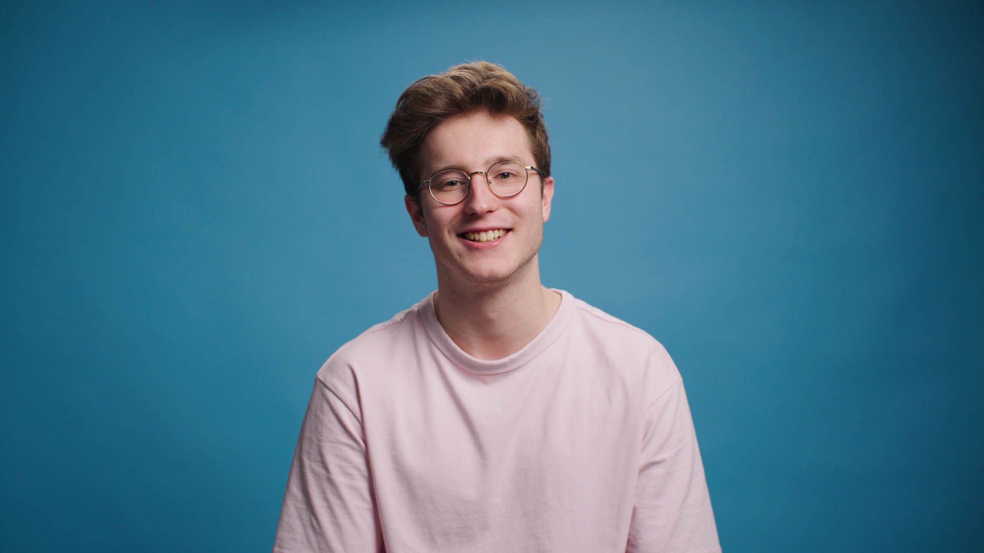 A young man wearing glasses and a pink shirt is smiling against a blue background.