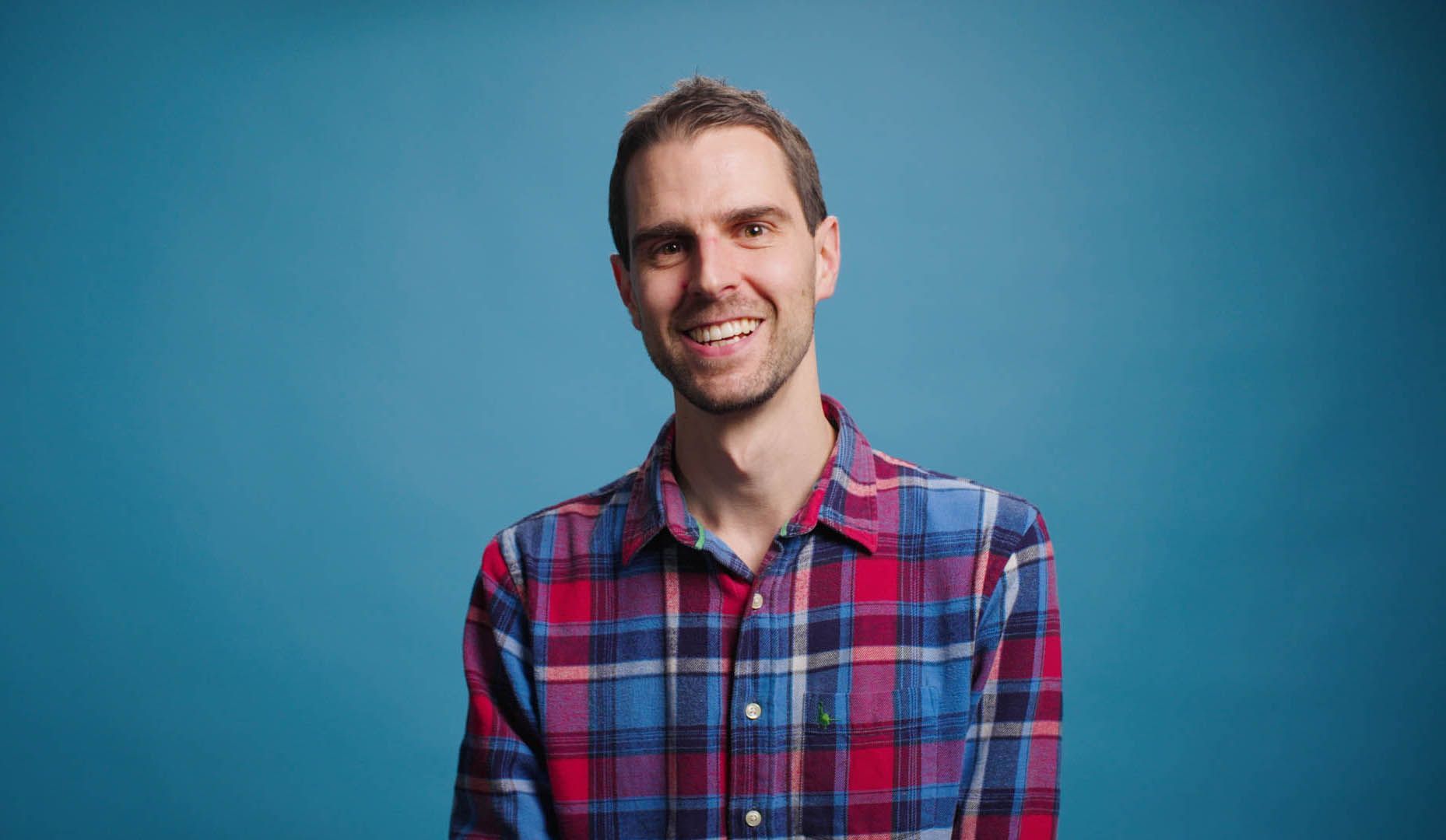 A man in a plaid shirt is smiling in front of a blue background.