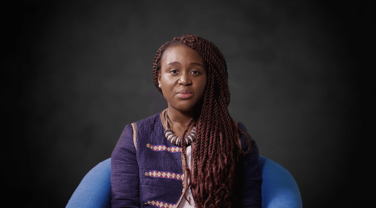 A woman with dreadlocks is sitting in a blue chair and looking at the camera.