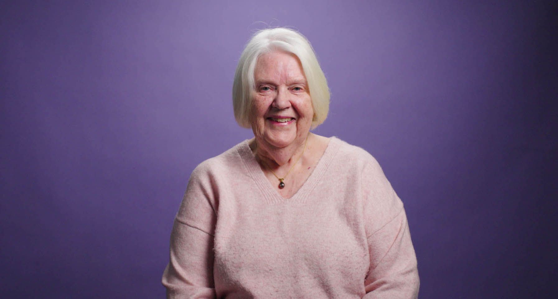 An elderly woman in a pink sweater is smiling in front of a purple background.