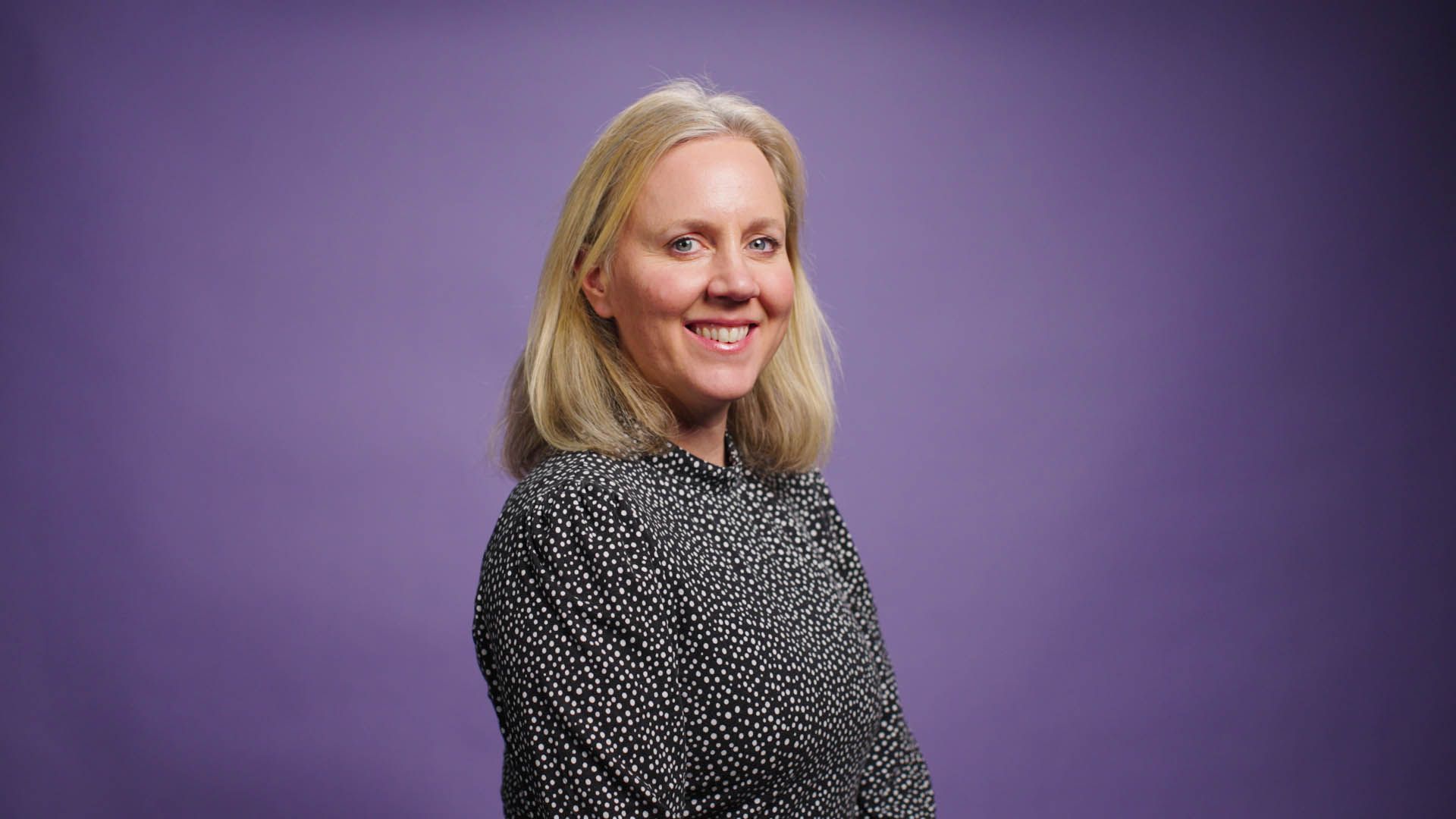 A woman is standing in front of a purple background and smiling.
