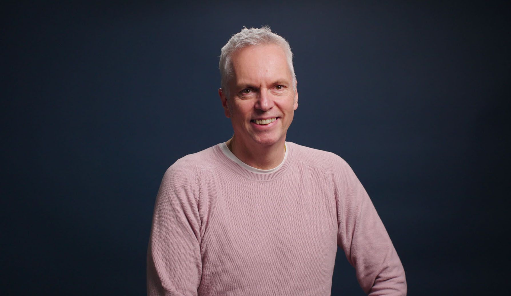 A man in a pink sweater is smiling in front of a blue background.