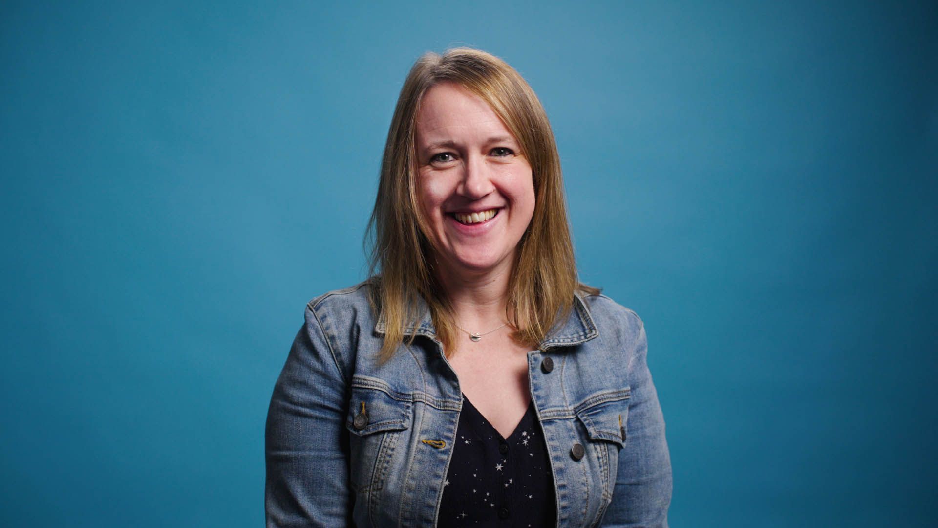 A woman in a denim jacket is smiling in front of a blue background.