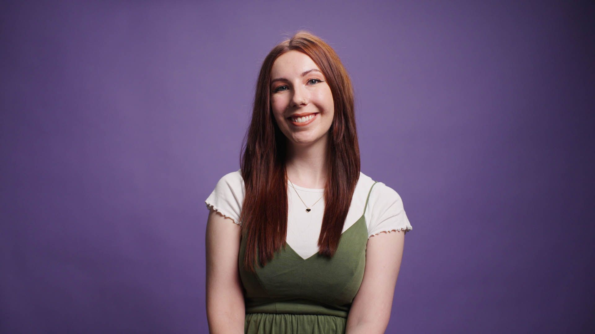 A woman with long red hair is smiling in front of a purple background.