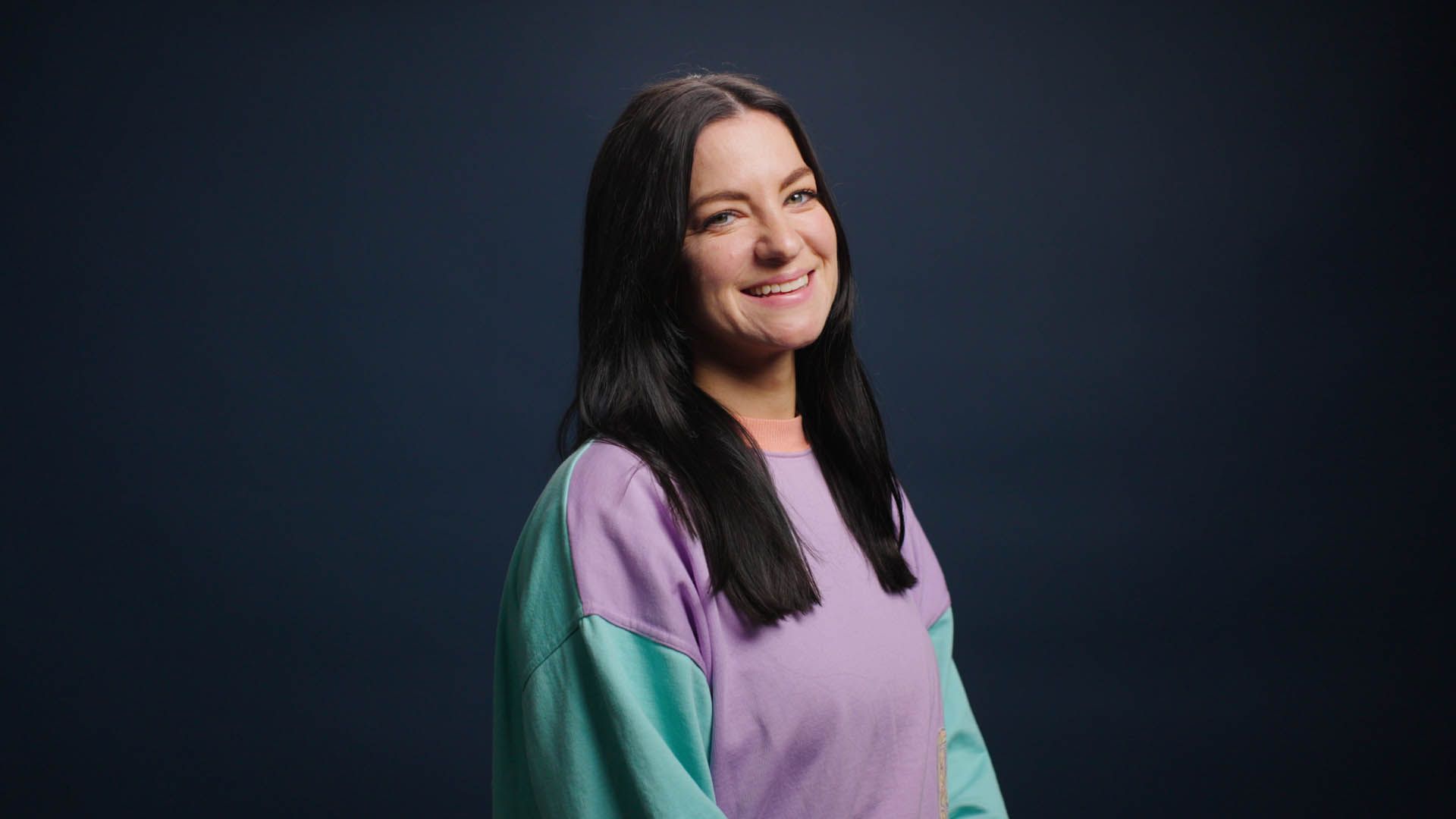 A woman in a purple and green sweater is smiling and looking at the camera.